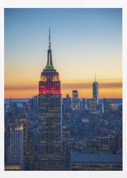 A vertical photograph of the New York City skyline at sunset, with the Empire State Building in the foreground. The building is illuminated with festive red and green lights on its upper floors, set against a sky that transitions from deep blue to a warm orange glow at the horizon. The surrounding cityscape, including the distant One World Trade Center, is bathed in the cool light of dusk, with scattered warm lights from building windows. Decor