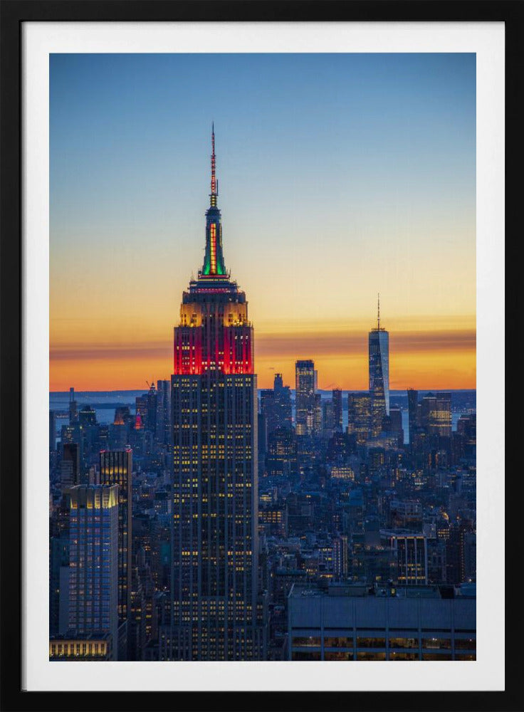 A vertical photograph of the New York City skyline at sunset, with the Empire State Building in the foreground. The building is illuminated with festive red and green lights on its upper floors, set against a sky that transitions from deep blue to a warm orange glow at the horizon. The surrounding cityscape, including the distant One World Trade Center, is bathed in the cool light of dusk, with scattered warm lights from building windows. Decor