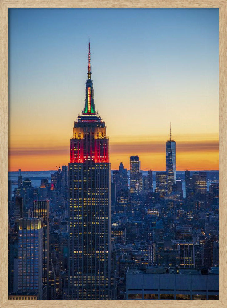 A vertical photograph of the New York City skyline at sunset, with the Empire State Building in the foreground. The building is illuminated with festive red and green lights on its upper floors, set against a sky that transitions from deep blue to a warm orange glow at the horizon. The surrounding cityscape, including the distant One World Trade Center, is bathed in the cool light of dusk, with scattered warm lights from building windows. Decor