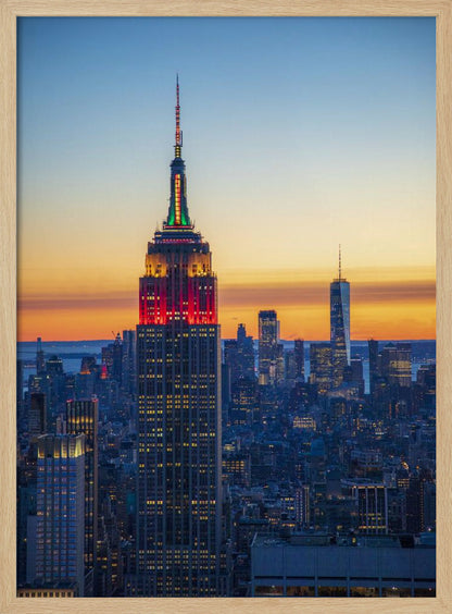 A vertical photograph of the New York City skyline at sunset, with the Empire State Building in the foreground. The building is illuminated with festive red and green lights on its upper floors, set against a sky that transitions from deep blue to a warm orange glow at the horizon. The surrounding cityscape, including the distant One World Trade Center, is bathed in the cool light of dusk, with scattered warm lights from building windows. Decor