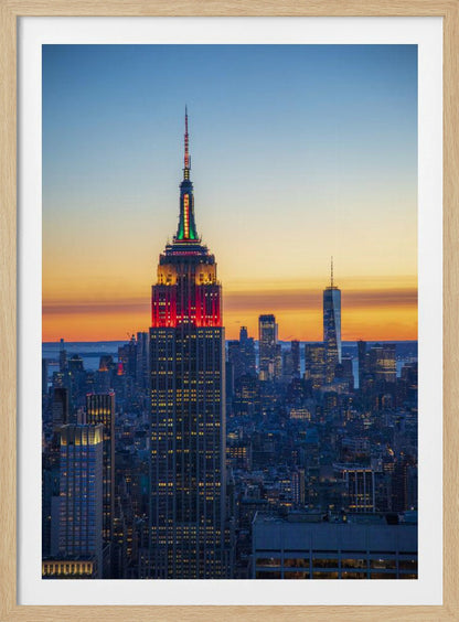 A vertical photograph of the New York City skyline at sunset, with the Empire State Building in the foreground. The building is illuminated with festive red and green lights on its upper floors, set against a sky that transitions from deep blue to a warm orange glow at the horizon. The surrounding cityscape, including the distant One World Trade Center, is bathed in the cool light of dusk, with scattered warm lights from building windows. Decor