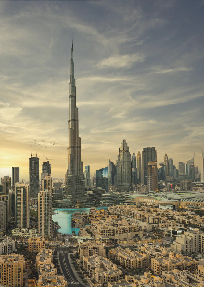 A framed photograph of the Dubai skyline at sunset, with the iconic Burj Khalifa skyscraper dominating the center. Surrounding it are numerous other modern high-rise buildings, a body of turquoise water, and a dense urban landscape in the foreground, all under a cloudy, golden-hued sky. Poster