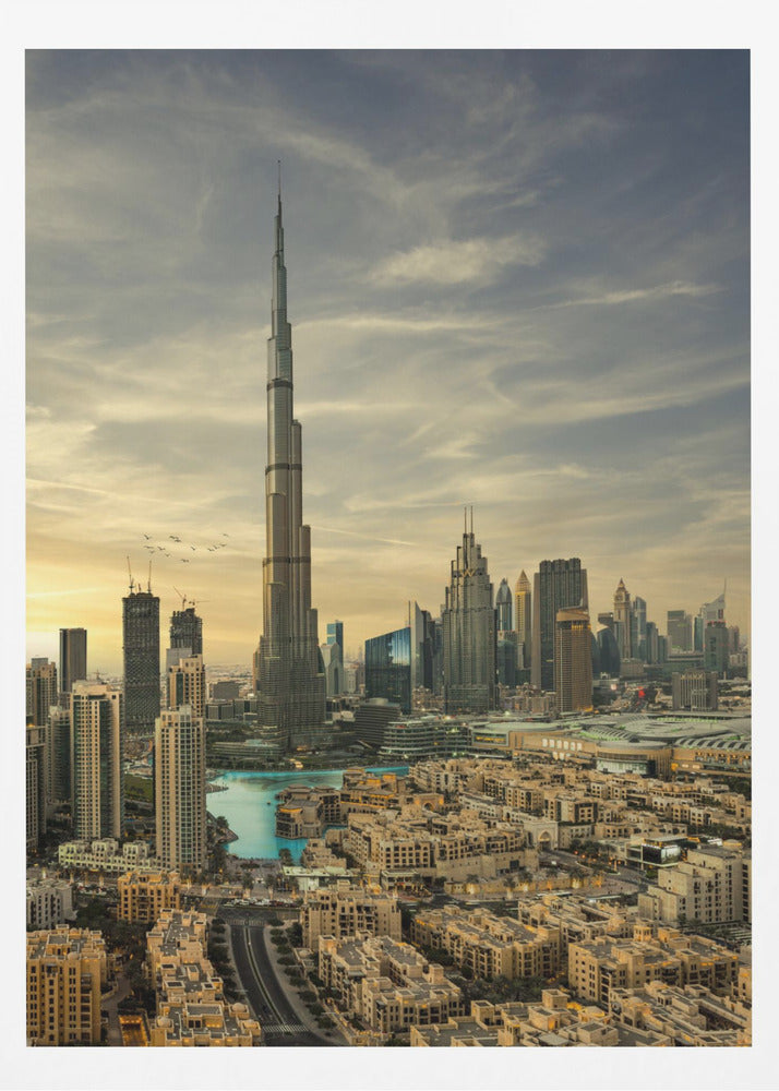 A framed photograph of the Dubai skyline at sunset, with the iconic Burj Khalifa skyscraper dominating the center. Surrounding it are numerous other modern high-rise buildings, a body of turquoise water, and a dense urban landscape in the foreground, all under a cloudy, golden-hued sky. Poster