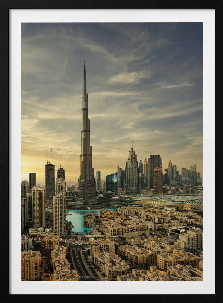 A framed photograph of the Dubai skyline at sunset, with the iconic Burj Khalifa skyscraper dominating the center. Surrounding it are numerous other modern high-rise buildings, a body of turquoise water, and a dense urban landscape in the foreground, all under a cloudy, golden-hued sky. Poster