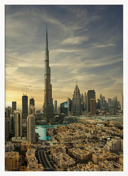 A framed photograph of the Dubai skyline at sunset, with the iconic Burj Khalifa skyscraper dominating the center. Surrounding it are numerous other modern high-rise buildings, a body of turquoise water, and a dense urban landscape in the foreground, all under a cloudy, golden-hued sky. Poster