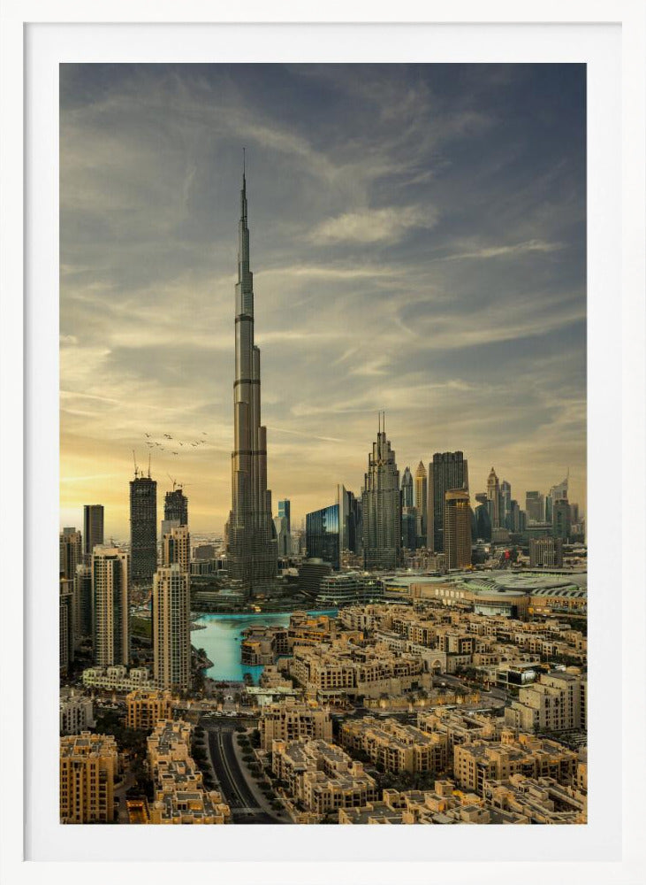 A framed photograph of the Dubai skyline at sunset, with the iconic Burj Khalifa skyscraper dominating the center. Surrounding it are numerous other modern high-rise buildings, a body of turquoise water, and a dense urban landscape in the foreground, all under a cloudy, golden-hued sky. Poster