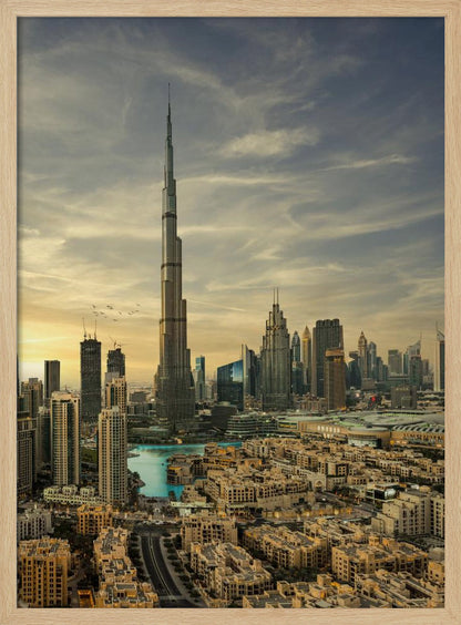 A framed photograph of the Dubai skyline at sunset, with the iconic Burj Khalifa skyscraper dominating the center. Surrounding it are numerous other modern high-rise buildings, a body of turquoise water, and a dense urban landscape in the foreground, all under a cloudy, golden-hued sky. Poster