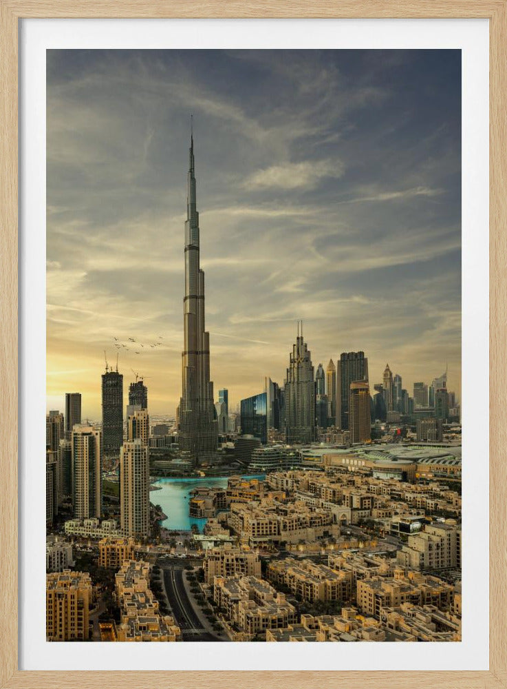 A framed photograph of the Dubai skyline at sunset, with the iconic Burj Khalifa skyscraper dominating the center. Surrounding it are numerous other modern high-rise buildings, a body of turquoise water, and a dense urban landscape in the foreground, all under a cloudy, golden-hued sky. Poster