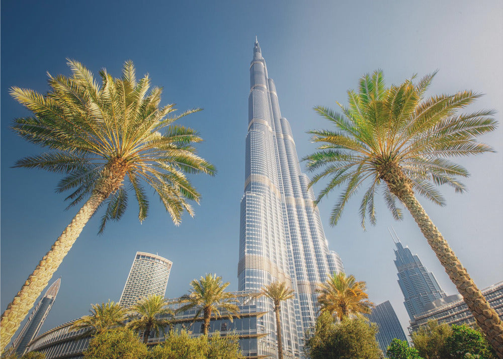 A low-angle photograph looking up at the towering Burj Khalifa skyscraper in Dubai, framed by two large palm trees against a clear, vibrant blue sky. Wall Art