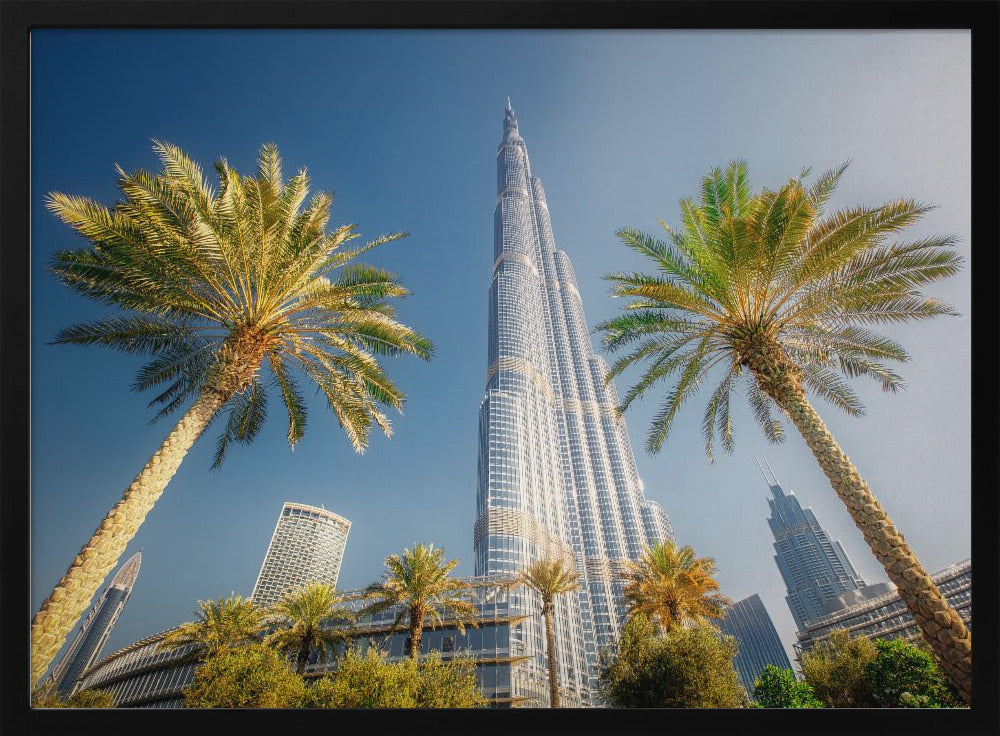 A low-angle photograph looking up at the towering Burj Khalifa skyscraper in Dubai, framed by two large palm trees against a clear, vibrant blue sky. Wall Art