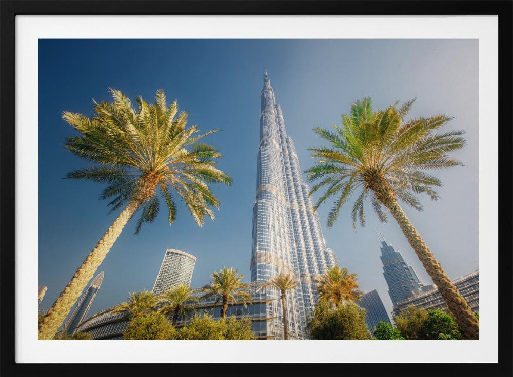 A low-angle photograph looking up at the towering Burj Khalifa skyscraper in Dubai, framed by two large palm trees against a clear, vibrant blue sky. Wall Art