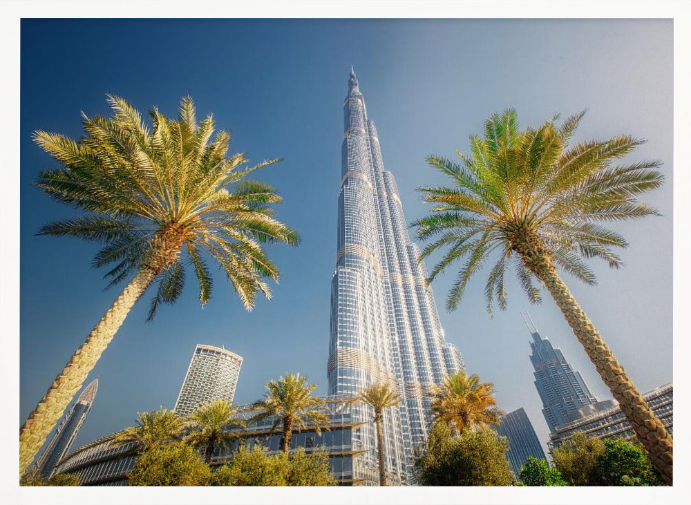 A low-angle photograph looking up at the towering Burj Khalifa skyscraper in Dubai, framed by two large palm trees against a clear, vibrant blue sky. Wall Art