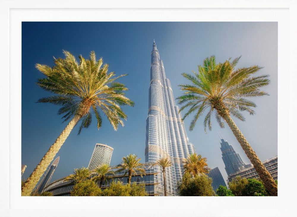 A low-angle photograph looking up at the towering Burj Khalifa skyscraper in Dubai, framed by two large palm trees against a clear, vibrant blue sky. Wall Art