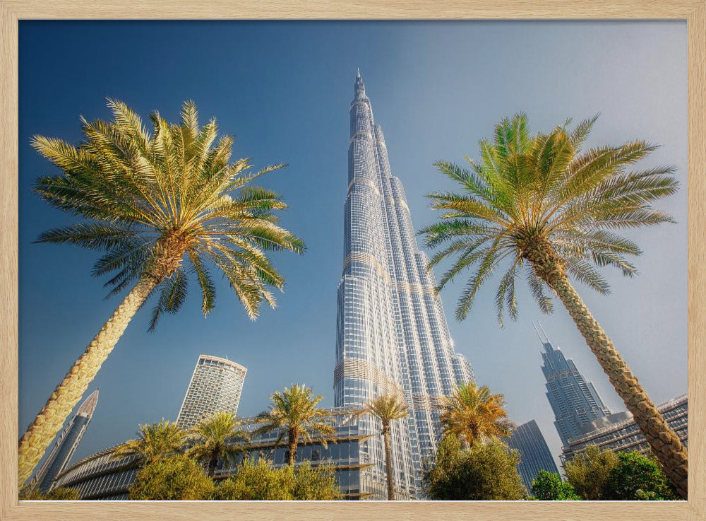 A low-angle photograph looking up at the towering Burj Khalifa skyscraper in Dubai, framed by two large palm trees against a clear, vibrant blue sky. Wall Art