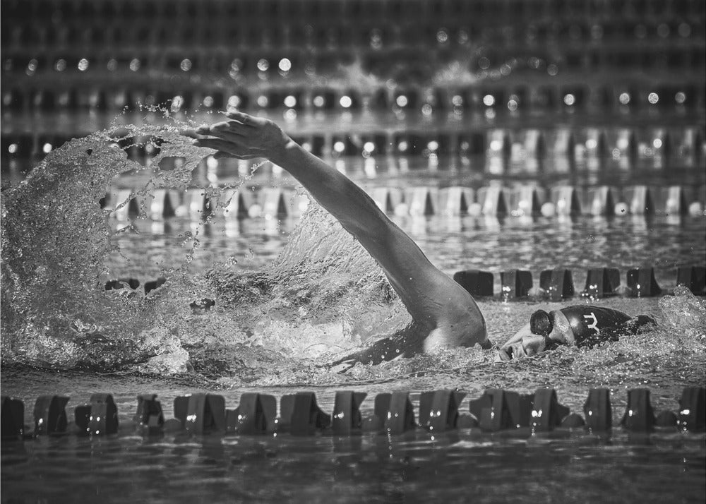 A dramatic black and white action photograph of a competitive swimmer mid-stroke, captured within a silver frame. The swimmer's arm is extended forward, creating a powerful splash of water. The focus is on the motion and intensity of the sport, with the pool's lane lines blurred in the background. Wall Art