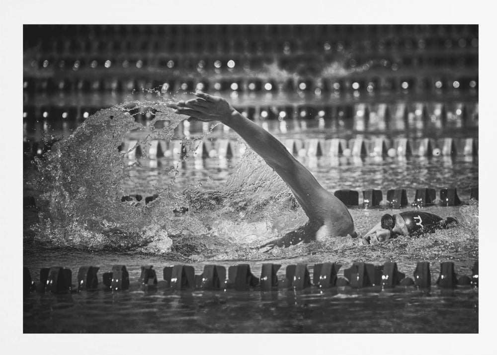 A dramatic black and white action photograph of a competitive swimmer mid-stroke, captured within a silver frame. The swimmer's arm is extended forward, creating a powerful splash of water. The focus is on the motion and intensity of the sport, with the pool's lane lines blurred in the background. Wall Art