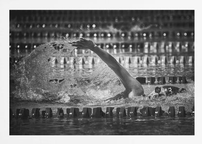 A dramatic black and white action photograph of a competitive swimmer mid-stroke, captured within a silver frame. The swimmer's arm is extended forward, creating a powerful splash of water. The focus is on the motion and intensity of the sport, with the pool's lane lines blurred in the background. Wall Art