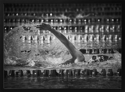 A dramatic black and white action photograph of a competitive swimmer mid-stroke, captured within a silver frame. The swimmer's arm is extended forward, creating a powerful splash of water. The focus is on the motion and intensity of the sport, with the pool's lane lines blurred in the background. Wall Art