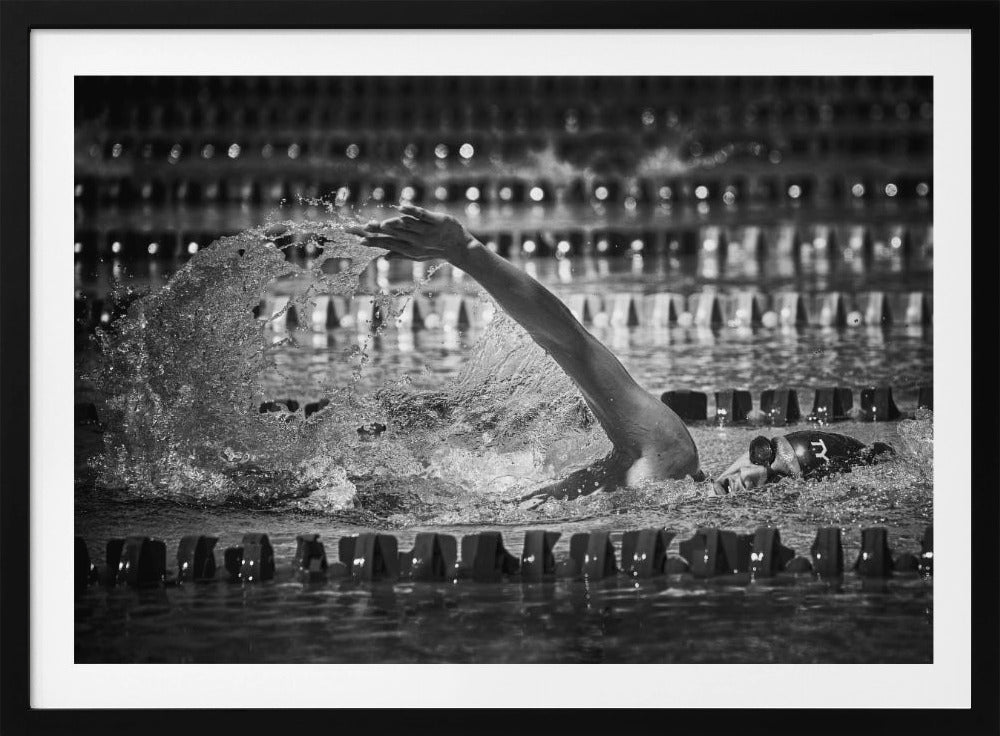A dramatic black and white action photograph of a competitive swimmer mid-stroke, captured within a silver frame. The swimmer's arm is extended forward, creating a powerful splash of water. The focus is on the motion and intensity of the sport, with the pool's lane lines blurred in the background. Wall Art
