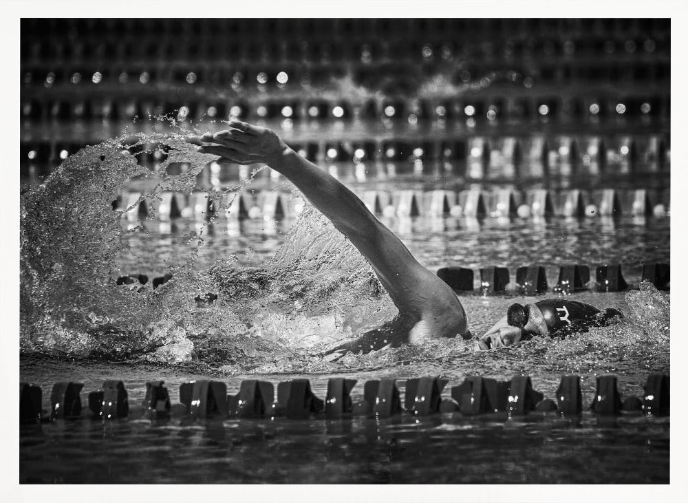 A dramatic black and white action photograph of a competitive swimmer mid-stroke, captured within a silver frame. The swimmer's arm is extended forward, creating a powerful splash of water. The focus is on the motion and intensity of the sport, with the pool's lane lines blurred in the background. Wall Art
