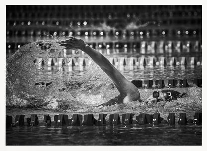 A dramatic black and white action photograph of a competitive swimmer mid-stroke, captured within a silver frame. The swimmer's arm is extended forward, creating a powerful splash of water. The focus is on the motion and intensity of the sport, with the pool's lane lines blurred in the background. Wall Art