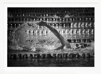 A dramatic black and white action photograph of a competitive swimmer mid-stroke, captured within a silver frame. The swimmer's arm is extended forward, creating a powerful splash of water. The focus is on the motion and intensity of the sport, with the pool's lane lines blurred in the background. Wall Art