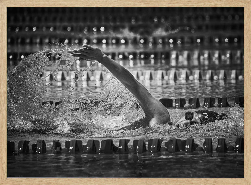 A dramatic black and white action photograph of a competitive swimmer mid-stroke, captured within a silver frame. The swimmer's arm is extended forward, creating a powerful splash of water. The focus is on the motion and intensity of the sport, with the pool's lane lines blurred in the background. Wall Art