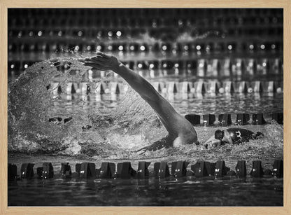 A dramatic black and white action photograph of a competitive swimmer mid-stroke, captured within a silver frame. The swimmer's arm is extended forward, creating a powerful splash of water. The focus is on the motion and intensity of the sport, with the pool's lane lines blurred in the background. Wall Art
