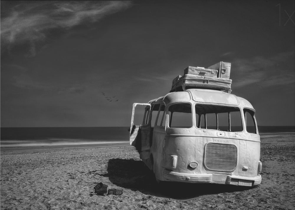 A dramatic black and white photograph of a vintage bus with luggage stacked on the roof, parked on a sandy beach. The ocean stretches out to the horizon under a dark, moody sky, with a small flock of birds flying in the distance. Print