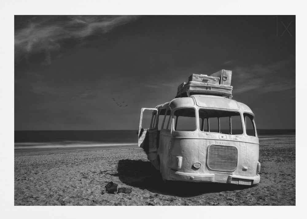 A dramatic black and white photograph of a vintage bus with luggage stacked on the roof, parked on a sandy beach. The ocean stretches out to the horizon under a dark, moody sky, with a small flock of birds flying in the distance. Print