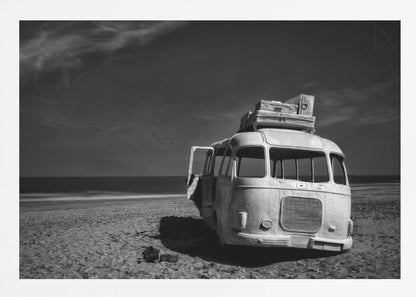 A dramatic black and white photograph of a vintage bus with luggage stacked on the roof, parked on a sandy beach. The ocean stretches out to the horizon under a dark, moody sky, with a small flock of birds flying in the distance. Print