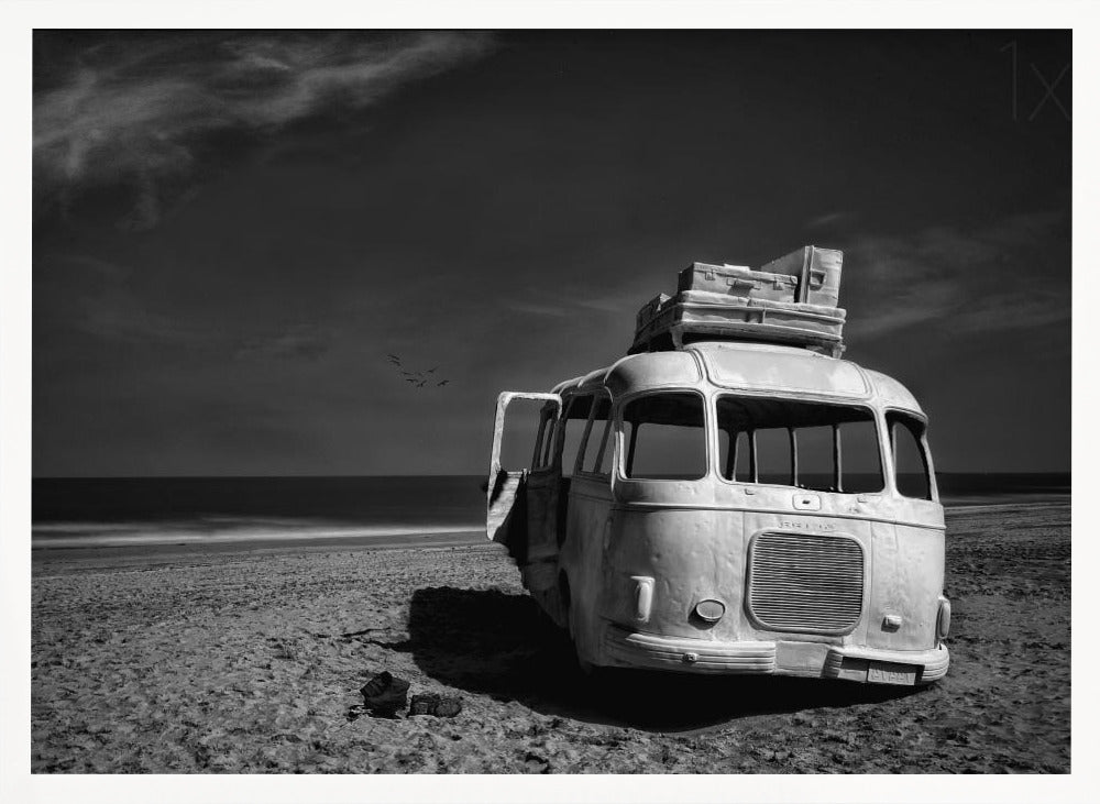 A dramatic black and white photograph of a vintage bus with luggage stacked on the roof, parked on a sandy beach. The ocean stretches out to the horizon under a dark, moody sky, with a small flock of birds flying in the distance. Print
