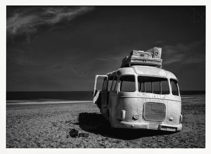 A dramatic black and white photograph of a vintage bus with luggage stacked on the roof, parked on a sandy beach. The ocean stretches out to the horizon under a dark, moody sky, with a small flock of birds flying in the distance. Print
