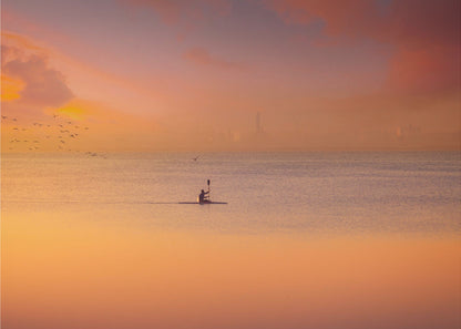 A serene photograph of a lone kayaker silhouetted against a vast expanse of calm water during a hazy, golden sunset. The sky and water are bathed in warm hues of orange, pink, and purple. A flock of birds flies in the distance, and the faint outline of a city skyline is visible on the horizon. Wall Art