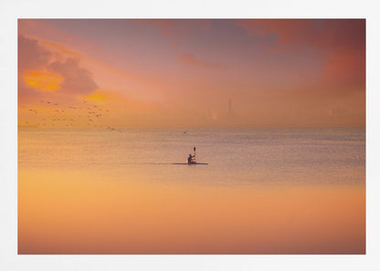 A serene photograph of a lone kayaker silhouetted against a vast expanse of calm water during a hazy, golden sunset. The sky and water are bathed in warm hues of orange, pink, and purple. A flock of birds flies in the distance, and the faint outline of a city skyline is visible on the horizon. Wall Art