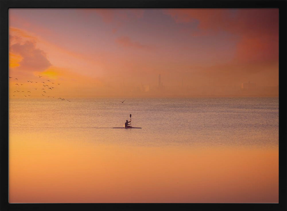 A serene photograph of a lone kayaker silhouetted against a vast expanse of calm water during a hazy, golden sunset. The sky and water are bathed in warm hues of orange, pink, and purple. A flock of birds flies in the distance, and the faint outline of a city skyline is visible on the horizon. Wall Art
