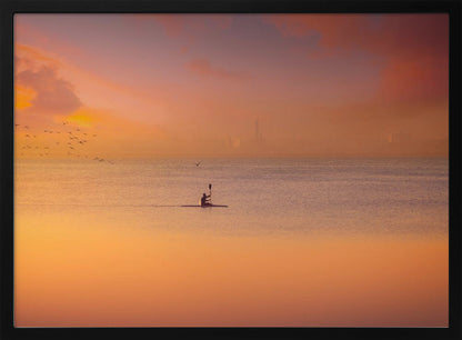 A serene photograph of a lone kayaker silhouetted against a vast expanse of calm water during a hazy, golden sunset. The sky and water are bathed in warm hues of orange, pink, and purple. A flock of birds flies in the distance, and the faint outline of a city skyline is visible on the horizon. Wall Art