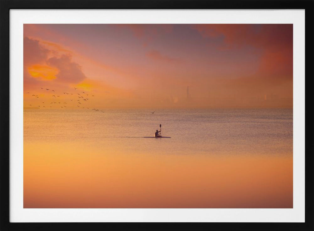A serene photograph of a lone kayaker silhouetted against a vast expanse of calm water during a hazy, golden sunset. The sky and water are bathed in warm hues of orange, pink, and purple. A flock of birds flies in the distance, and the faint outline of a city skyline is visible on the horizon. Wall Art