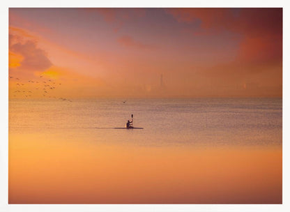 A serene photograph of a lone kayaker silhouetted against a vast expanse of calm water during a hazy, golden sunset. The sky and water are bathed in warm hues of orange, pink, and purple. A flock of birds flies in the distance, and the faint outline of a city skyline is visible on the horizon. Wall Art