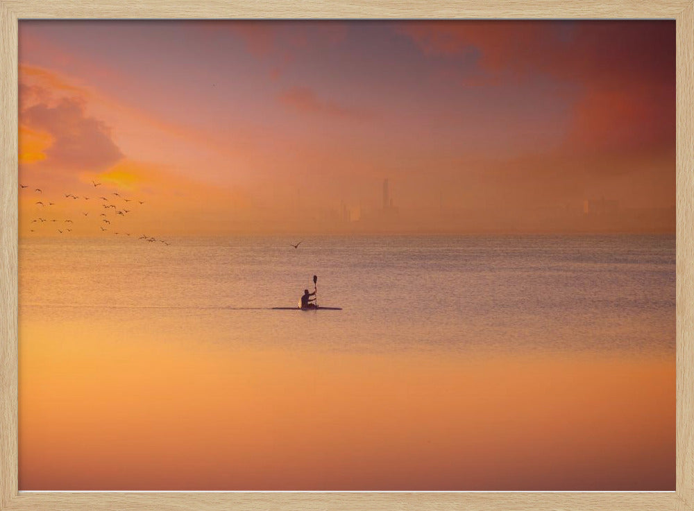 A serene photograph of a lone kayaker silhouetted against a vast expanse of calm water during a hazy, golden sunset. The sky and water are bathed in warm hues of orange, pink, and purple. A flock of birds flies in the distance, and the faint outline of a city skyline is visible on the horizon. Wall Art