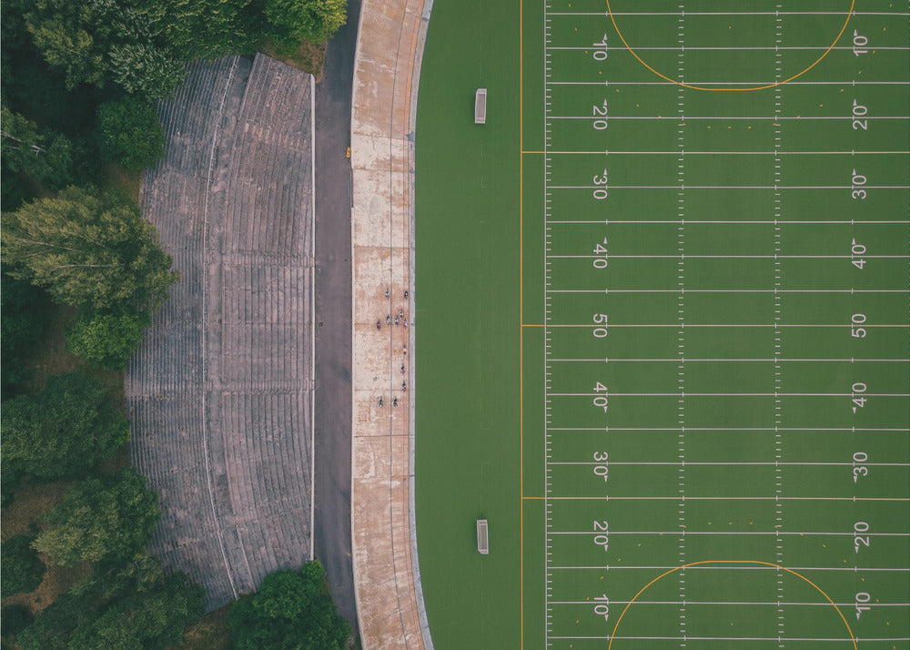 An aerial top-down photograph of an American football field beside empty, weathered concrete stadium bleachers. The vibrant green field is marked with white yard lines, and the gray stands are nestled amongst lush green trees. The entire scene is enclosed in a silver frame. Artwork