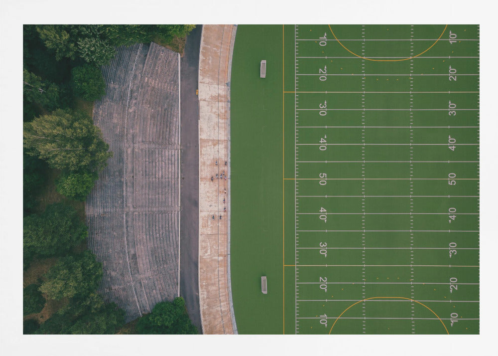 An aerial top-down photograph of an American football field beside empty, weathered concrete stadium bleachers. The vibrant green field is marked with white yard lines, and the gray stands are nestled amongst lush green trees. The entire scene is enclosed in a silver frame. Artwork