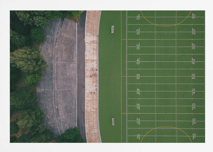 An aerial top-down photograph of an American football field beside empty, weathered concrete stadium bleachers. The vibrant green field is marked with white yard lines, and the gray stands are nestled amongst lush green trees. The entire scene is enclosed in a silver frame. Artwork