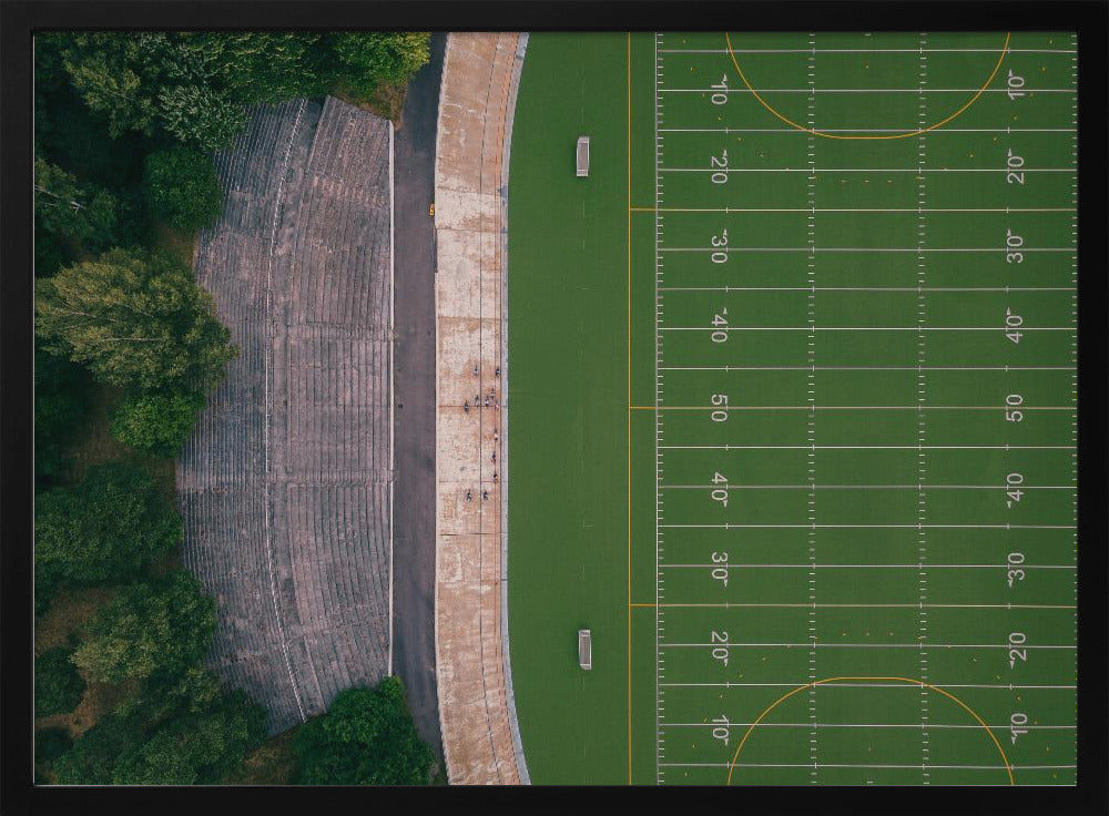 An aerial top-down photograph of an American football field beside empty, weathered concrete stadium bleachers. The vibrant green field is marked with white yard lines, and the gray stands are nestled amongst lush green trees. The entire scene is enclosed in a silver frame. Artwork