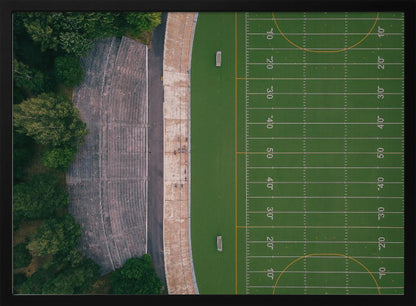 An aerial top-down photograph of an American football field beside empty, weathered concrete stadium bleachers. The vibrant green field is marked with white yard lines, and the gray stands are nestled amongst lush green trees. The entire scene is enclosed in a silver frame. Artwork