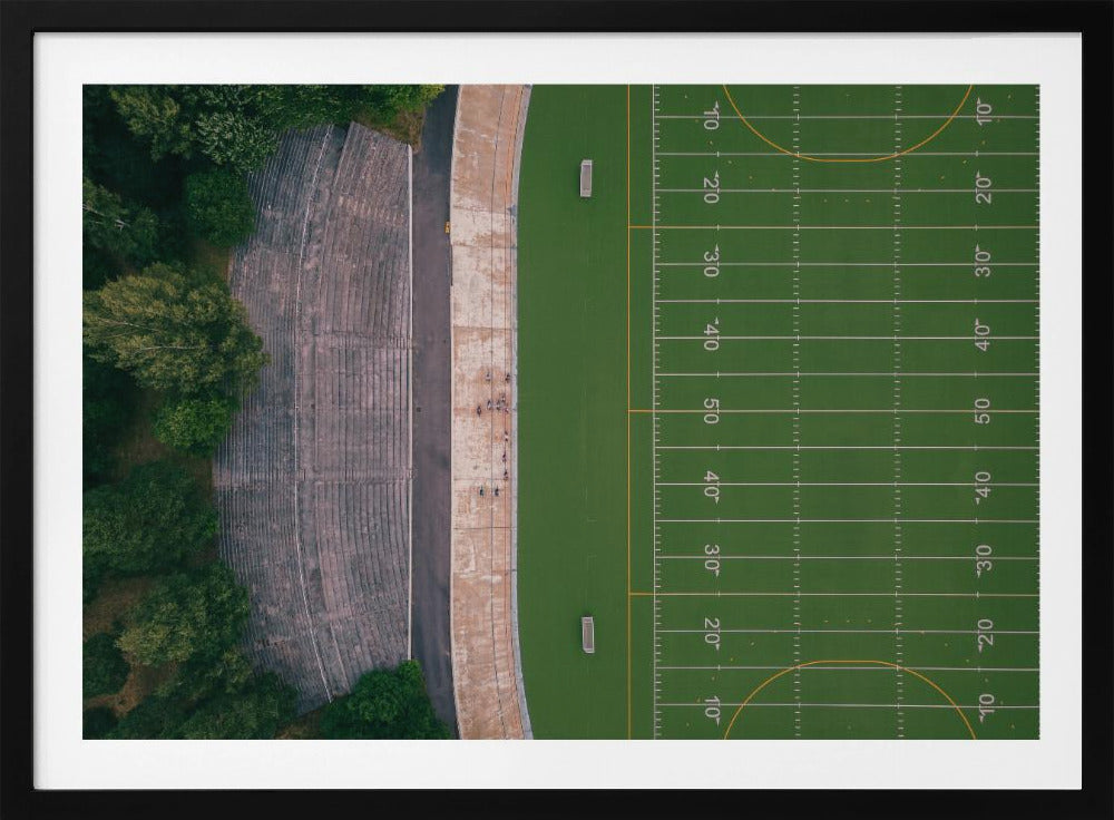 An aerial top-down photograph of an American football field beside empty, weathered concrete stadium bleachers. The vibrant green field is marked with white yard lines, and the gray stands are nestled amongst lush green trees. The entire scene is enclosed in a silver frame. Artwork