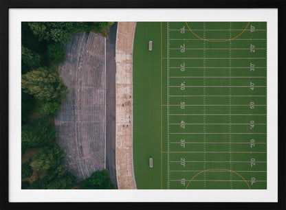 An aerial top-down photograph of an American football field beside empty, weathered concrete stadium bleachers. The vibrant green field is marked with white yard lines, and the gray stands are nestled amongst lush green trees. The entire scene is enclosed in a silver frame. Artwork