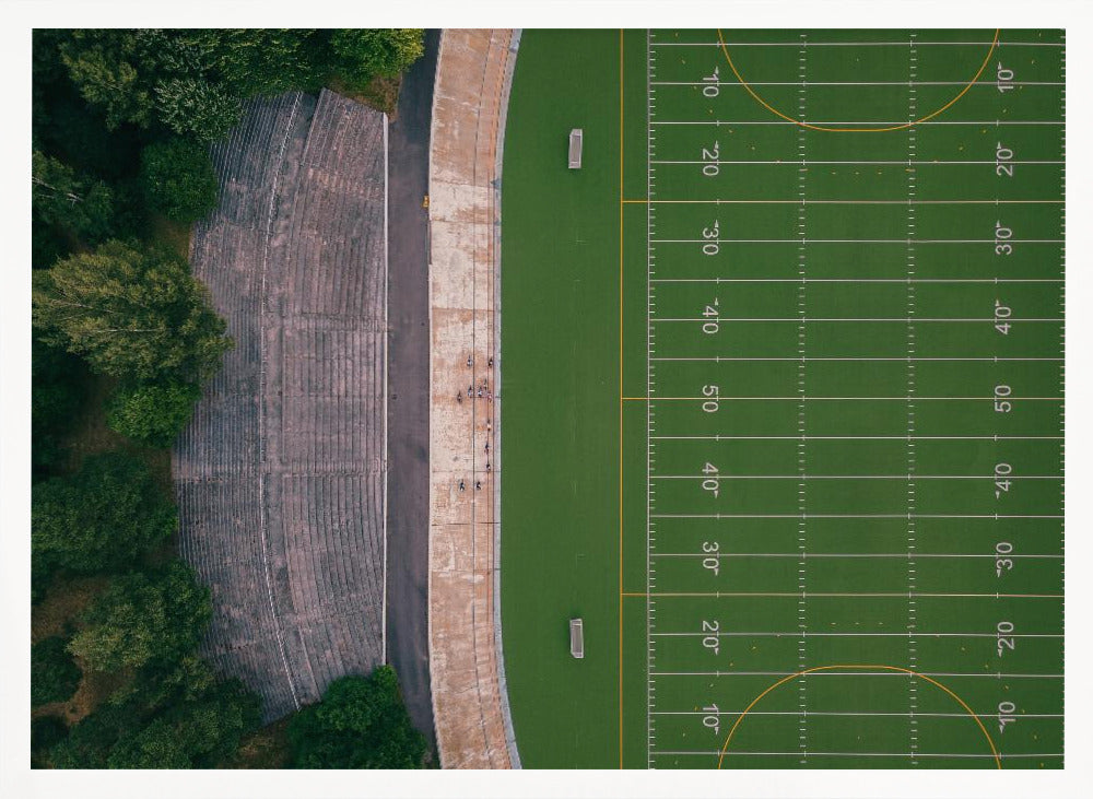 An aerial top-down photograph of an American football field beside empty, weathered concrete stadium bleachers. The vibrant green field is marked with white yard lines, and the gray stands are nestled amongst lush green trees. The entire scene is enclosed in a silver frame. Artwork