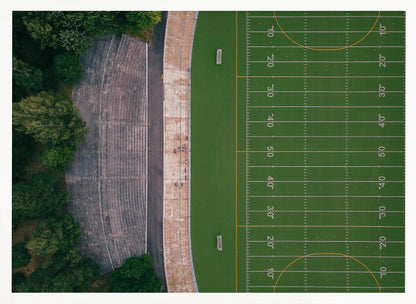 An aerial top-down photograph of an American football field beside empty, weathered concrete stadium bleachers. The vibrant green field is marked with white yard lines, and the gray stands are nestled amongst lush green trees. The entire scene is enclosed in a silver frame. Artwork