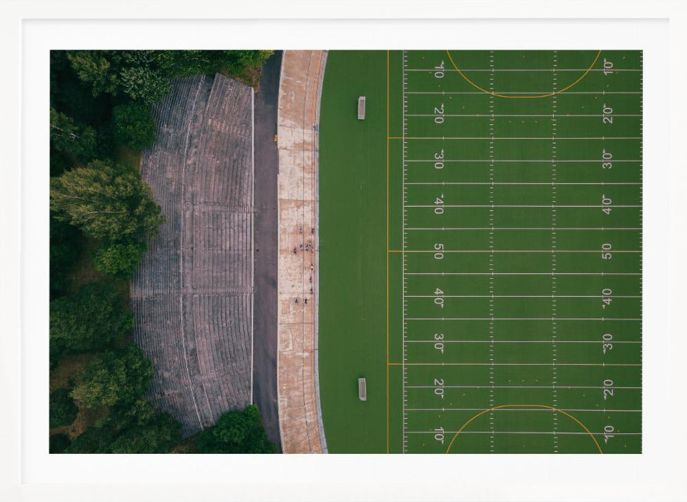 An aerial top-down photograph of an American football field beside empty, weathered concrete stadium bleachers. The vibrant green field is marked with white yard lines, and the gray stands are nestled amongst lush green trees. The entire scene is enclosed in a silver frame. Artwork